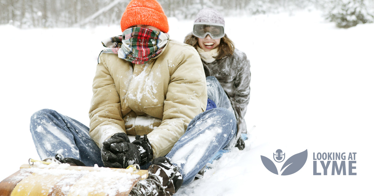 Two people cram themselves onto a toboggan in the deep snow with their toques and scarfs laughing and hanging out.