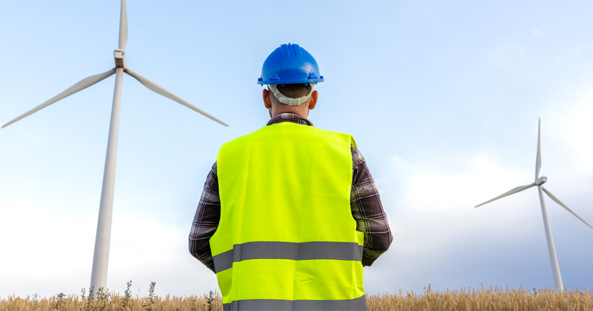 An outdoor engineer worker stands in front of giant wind powered electricity generators standing in a field of grass.