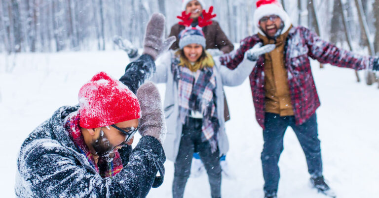 Friends and family play in the snow and attack their friend with snowballs, wearing Christmas hats.